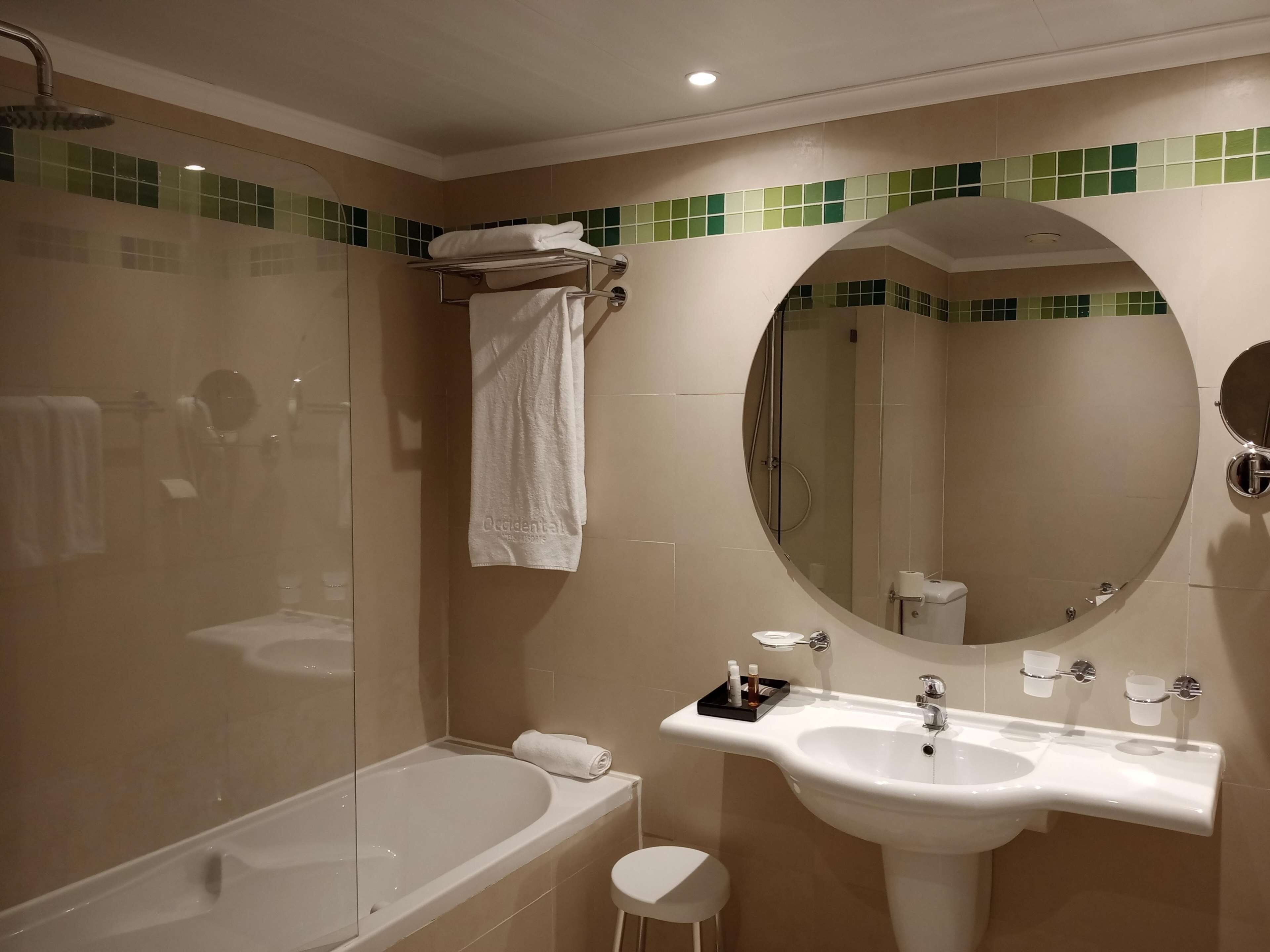 A modern bathroom with a bathtub, a round mirror, a sink with toiletries, and a glass-enclosed shower area.