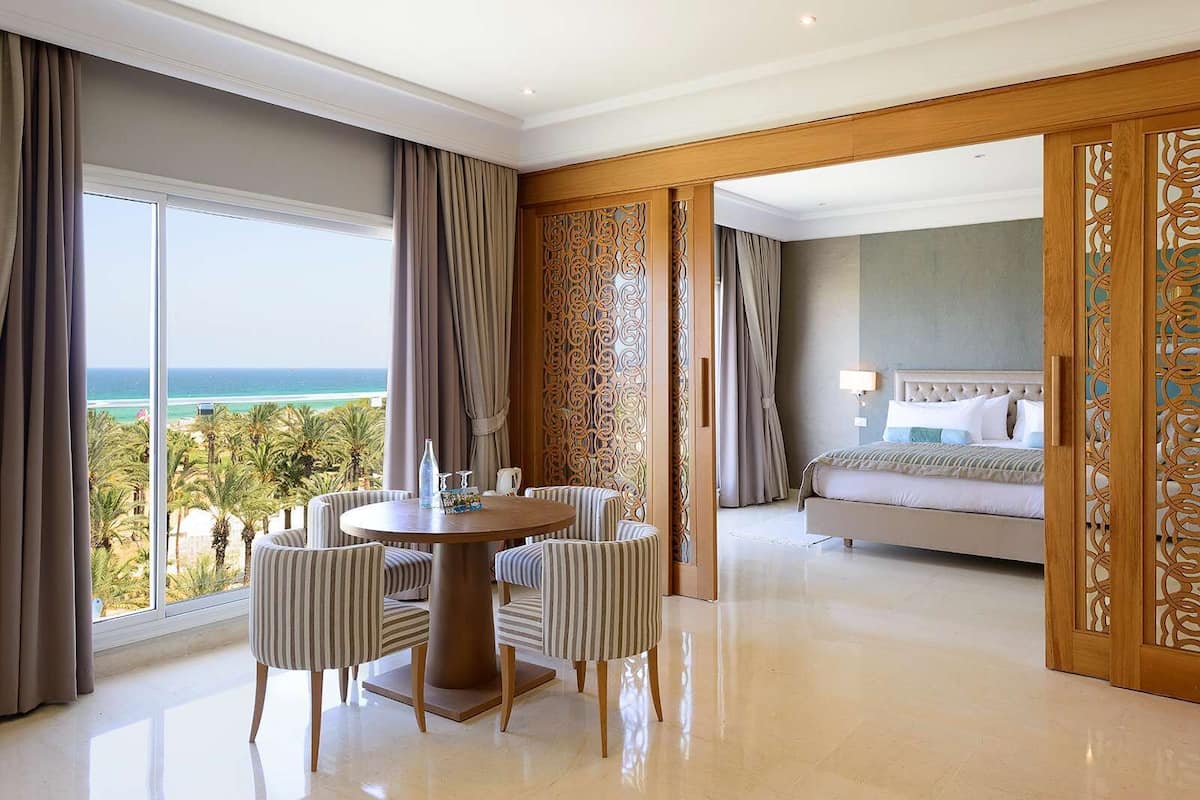 Une chambre d’hôtel dotée d’une grande fenêtre donnant sur l’océan, d’une table de salle à manger ronde avec des chaises à rayures et d’un lit avec une tête de lit.