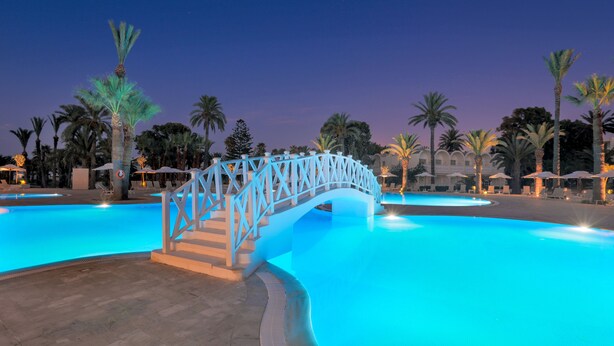 A swimming pool with a white bridge, palm trees, and a building in the background. - Occidental Sousse Marhaba (Sousse)