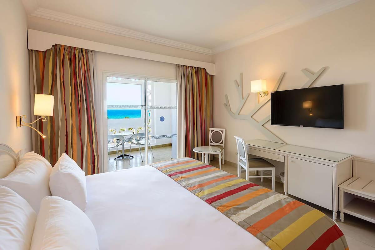 Une chambre d’hôtel avec un lit, un bureau, une chaise, une télévision et une vue sur l’océan par la fenêtre.