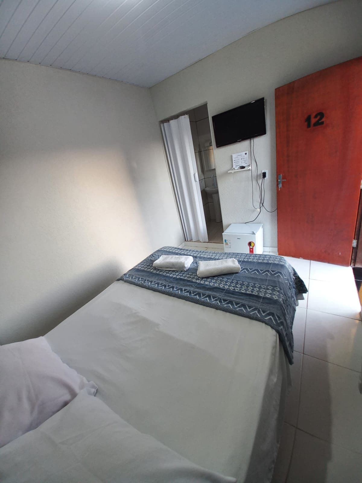 Een eenpersoonskamer met een bed, een nachtkastje, een aan de muur bevestigde televisie en een deur met het nummer 12.