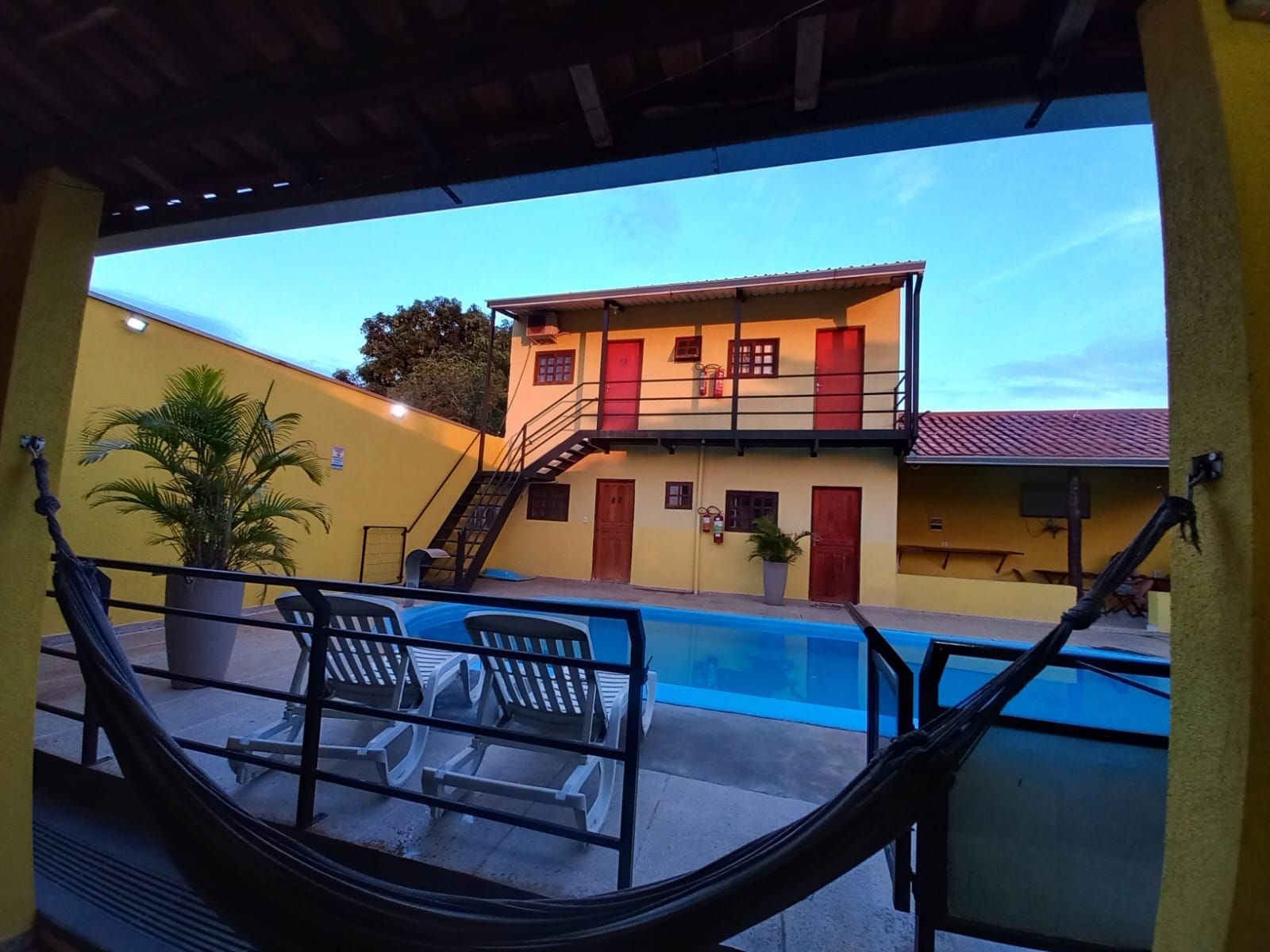 A pool area with lounge chairs and a view of a two-story building with a red door and a staircase.