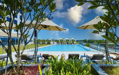 A swimming pool with lounge chairs and umbrellas, surrounded by greenery and a clear sky.