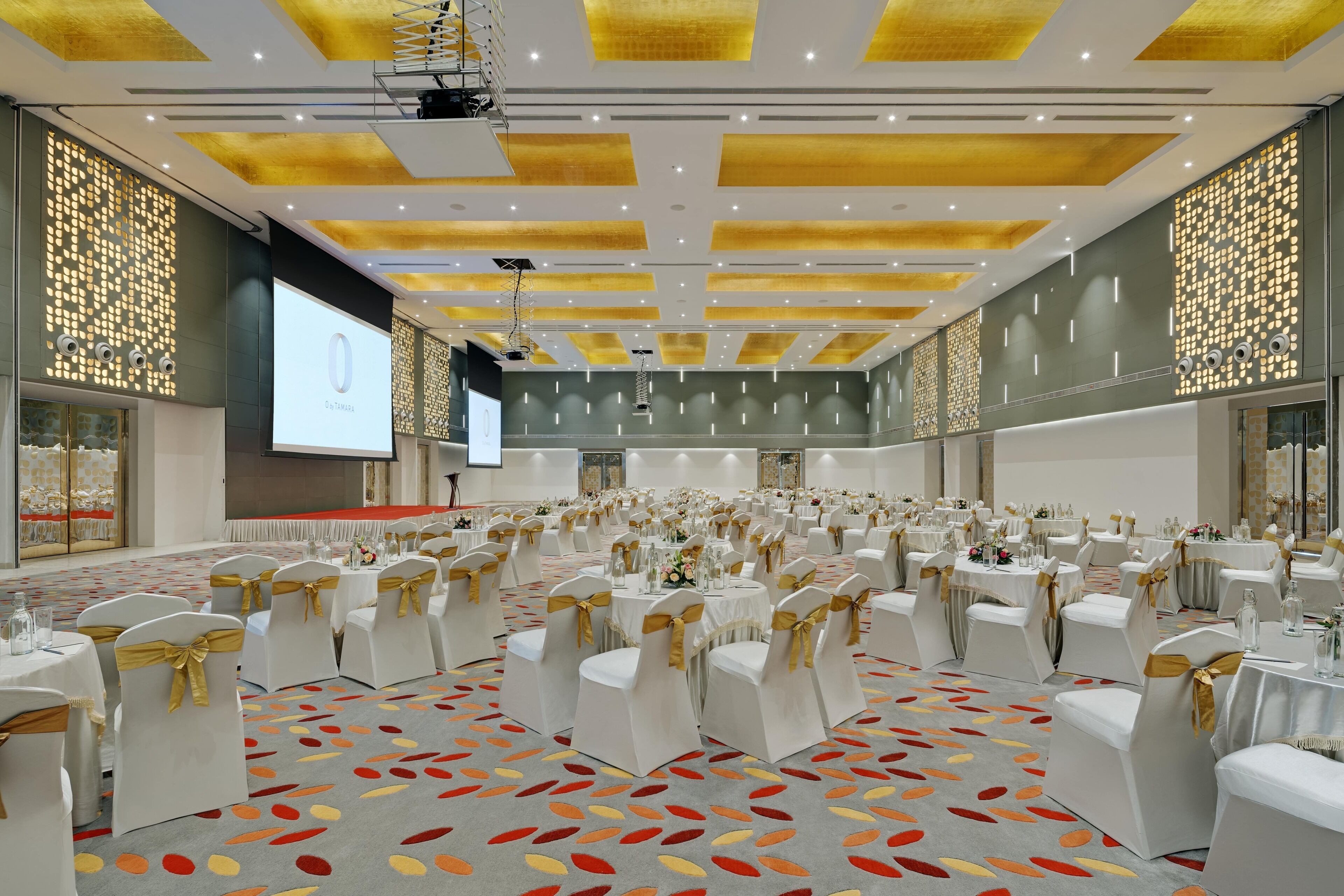 Un amplio salón de banquetes con filas de mesas y sillas, una pantalla de proyector y una alfombra estampada.