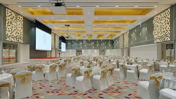 Une grande salle de banquet avec des rangées de tables et de chaises, un écran de projection et une moquette à motifs.