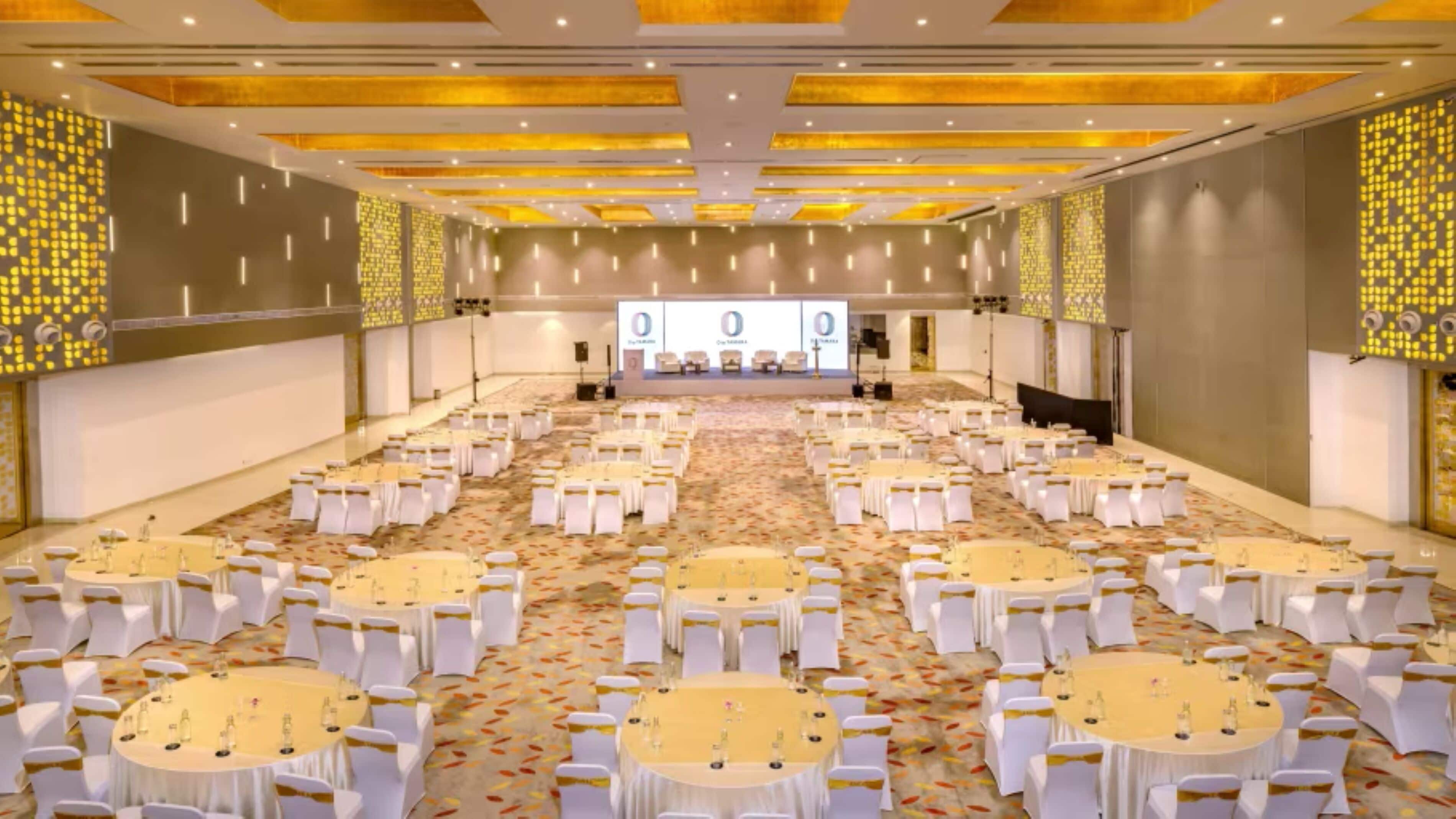 A large conference room with multiple round tables and white chairs, a stage with a podium, and patterned walls.