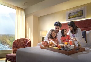 Un petit-déjeuner en famille, pris sur le lit d’un hôtel avec vue sur la ville, une chaise rouge à l’assise rembourrée et un plateau rempli de fruits et de boissons.