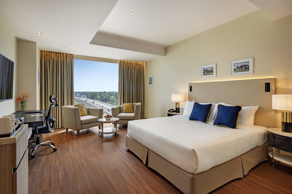A hotel room with a large bed, a desk with a chair, a sofa, and a view of a cityscape through the window.