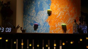 Una zona de bar con un reloj marcando las 23:08, una pared con arte abstracto colorido y botellas en la barra.