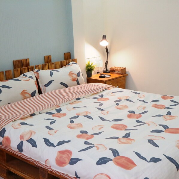 Una cama bien hecha con ropa de cama floral, una lámpara de noche y una mesita de madera pequeña con una planta y una cesta.