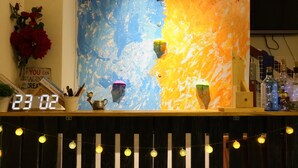 Un área de bar con un reloj marcando las 23:02, una pared con diseño abstracto azul y naranja, y luces de tipo guirnalda en el suelo.