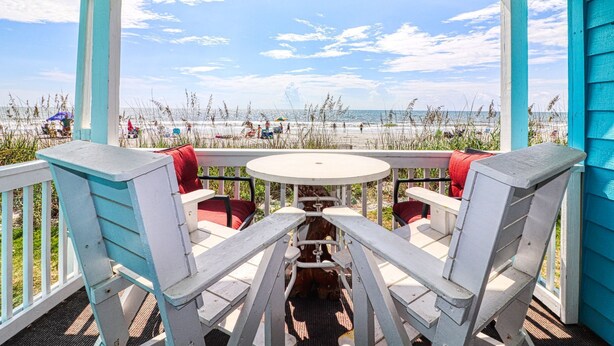 A covered outdoor seating area with a table and chairs, overlooking a beach with people and umbrellas. - Beach House 107 - Two Bedroom, Two Bath Oceanfront Condo (Garden City Beach)
