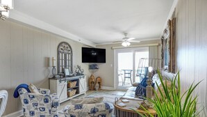 A living room with a television, a ceiling fan, a dining area with a table and chairs, and a large window. - Beach House 107 - Two Bedroom, Two Bath Oceanfront Condo (Garden City Beach)