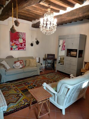 A cozy living room with a sofa, armchair, and a patterned rug. A chandelier hangs from the ceiling, and a cabinet is against the wall.