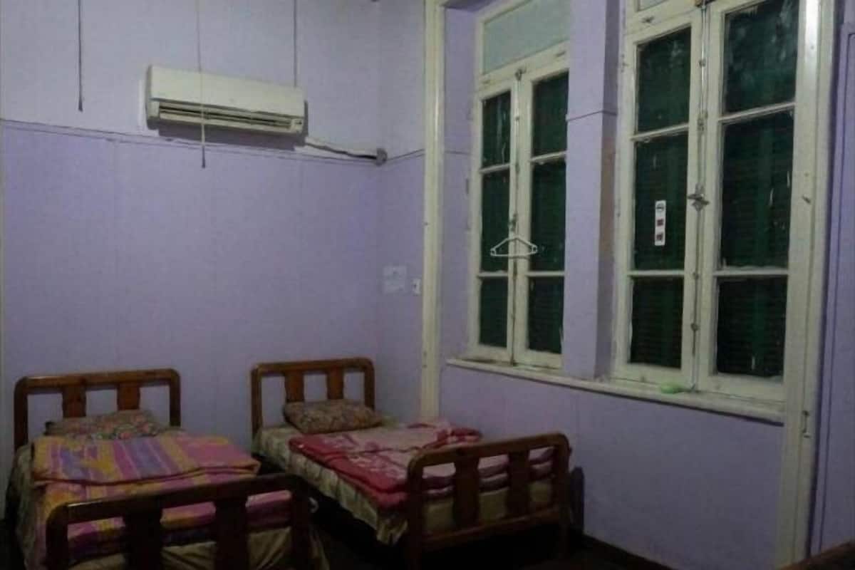 Une chambre avec deux lits en bois, une fenêtre avec des stores verts et un appareil de climatisation fixé au mur.