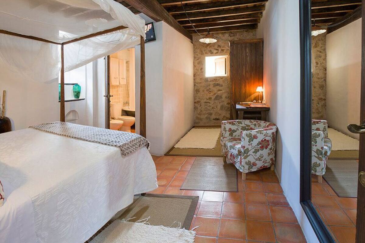A bedroom with a bed, a small table, and a chair. There is a bathroom visible through an open door.