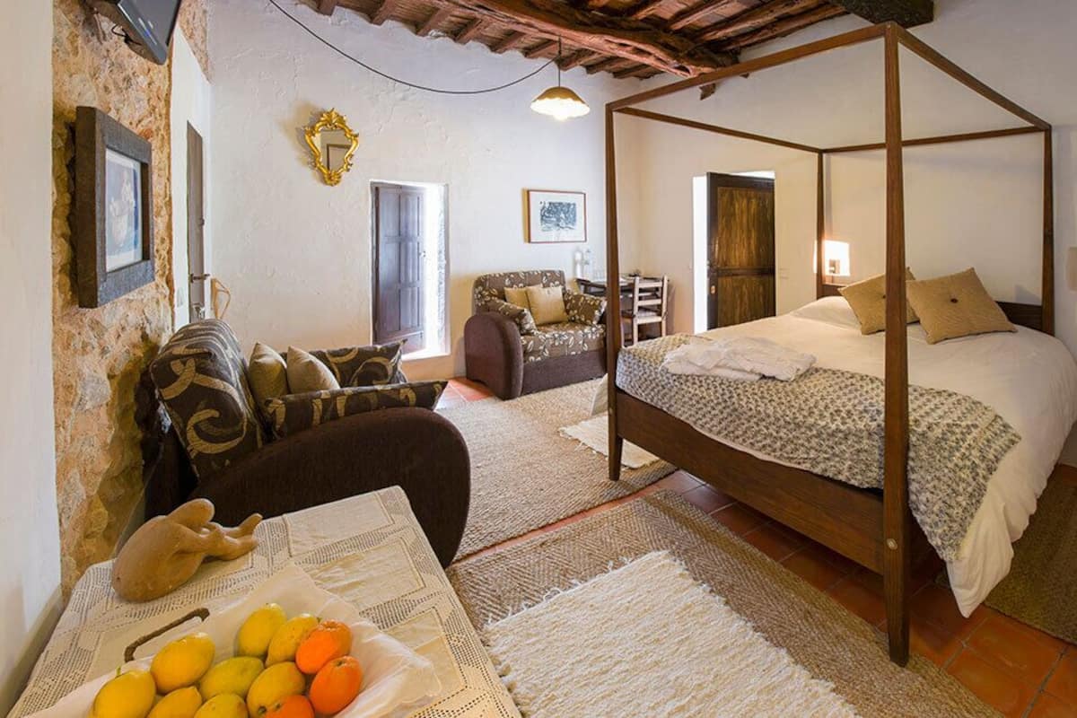 A bedroom with a four-poster bed, a sofa, a chair, and a table with fruit.