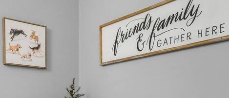 A hallway with a grey wall, a framed sign that reads 'friends family & gather here,' a small round table with a decorative item, and a framed picture of dogs.