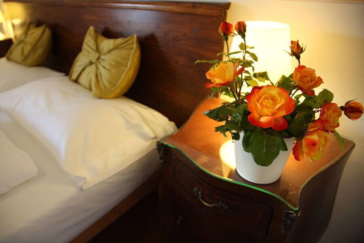 Un letto rifatto con cuscini, un comodino con un vaso di rose e una lampada.