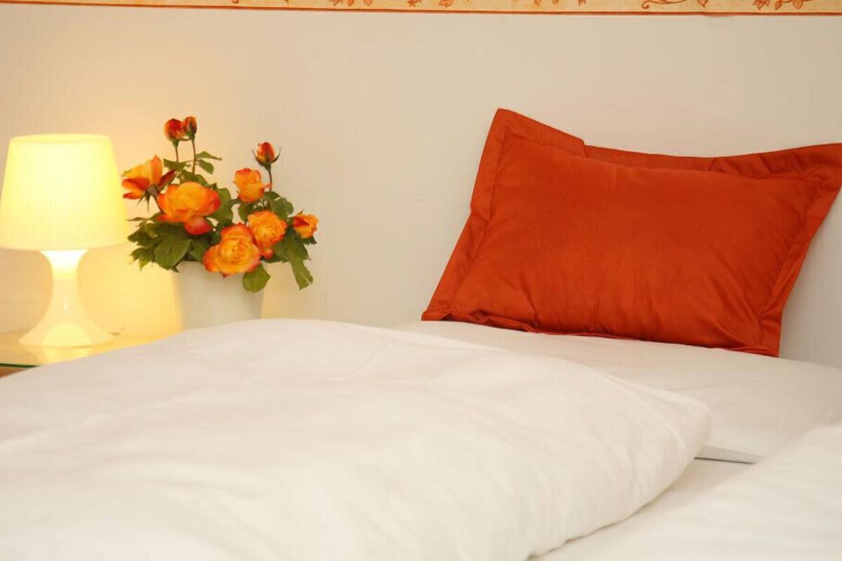 Un letto ben rifatto con lenzuola bianche, un cuscino arancione e un comodino con una lampada e un vaso di fiori arancioni.