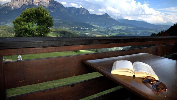Una panchina di legno con un libro aperto e occhiali, affacciata su un paesaggio montuoso.
