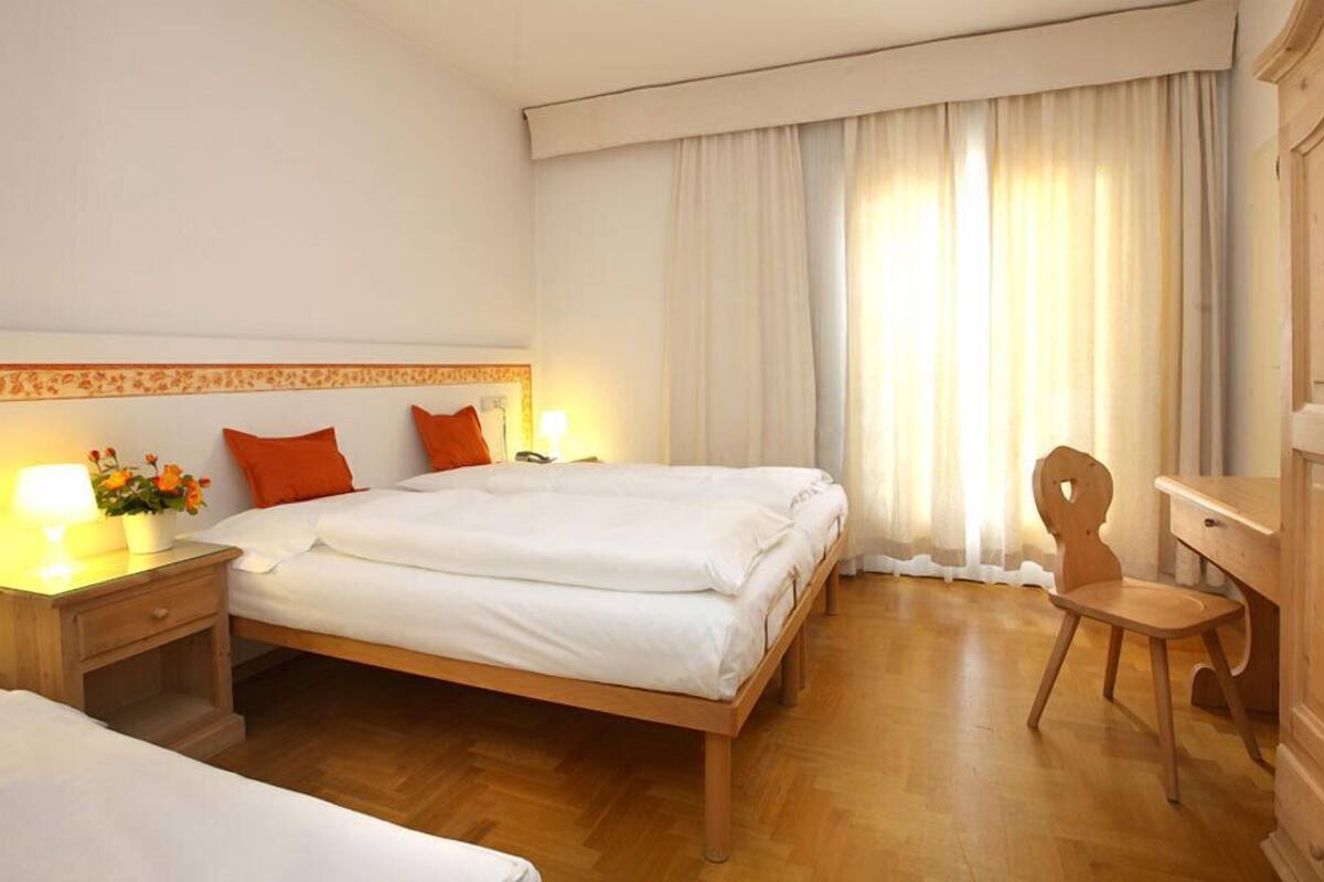 Camera d'albergo con un letto di legno, due cuscini, un comodino con una lampada, una sedia e un armadio.