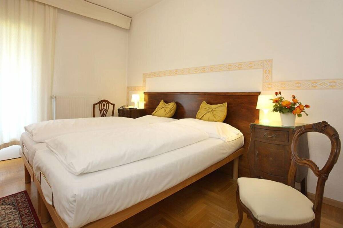 Una camera d'albergo con un letto grande, un comodino in legno, una sedia e un tavolino con una lampada e fiori.