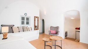 Un salon moderne avec un canapé blanc, un fauteuil gris, deux petites tables d’appoint en bois et un petit coin cuisine équipé d’un lave-vaisselle.