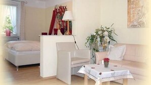 Une pièce comprenant un lit, un fauteuil blanc, une table recouverte d’une nappe, un vase avec des fleurs et un tableau accroché au mur.