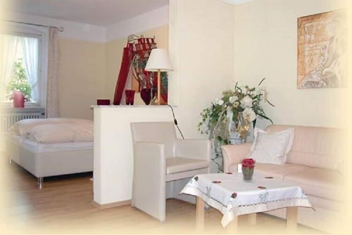 Una habitación con una cama, un sillón blanco, una mesa con mantel, un jarrón con flores y un cuadro en la pared.
