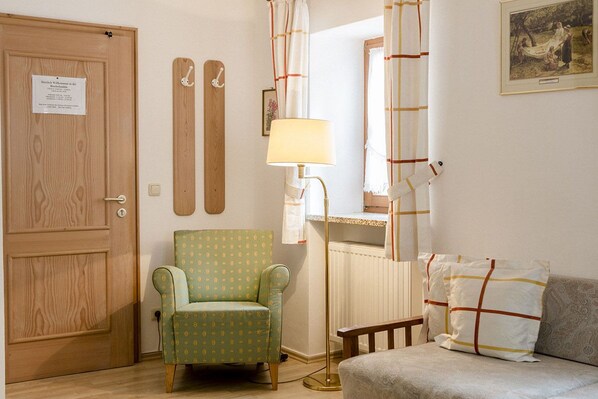 A room with a wooden door, a green patterned armchair, a floor lamp, a sofa with patterned cushions, and a framed picture on the wall.