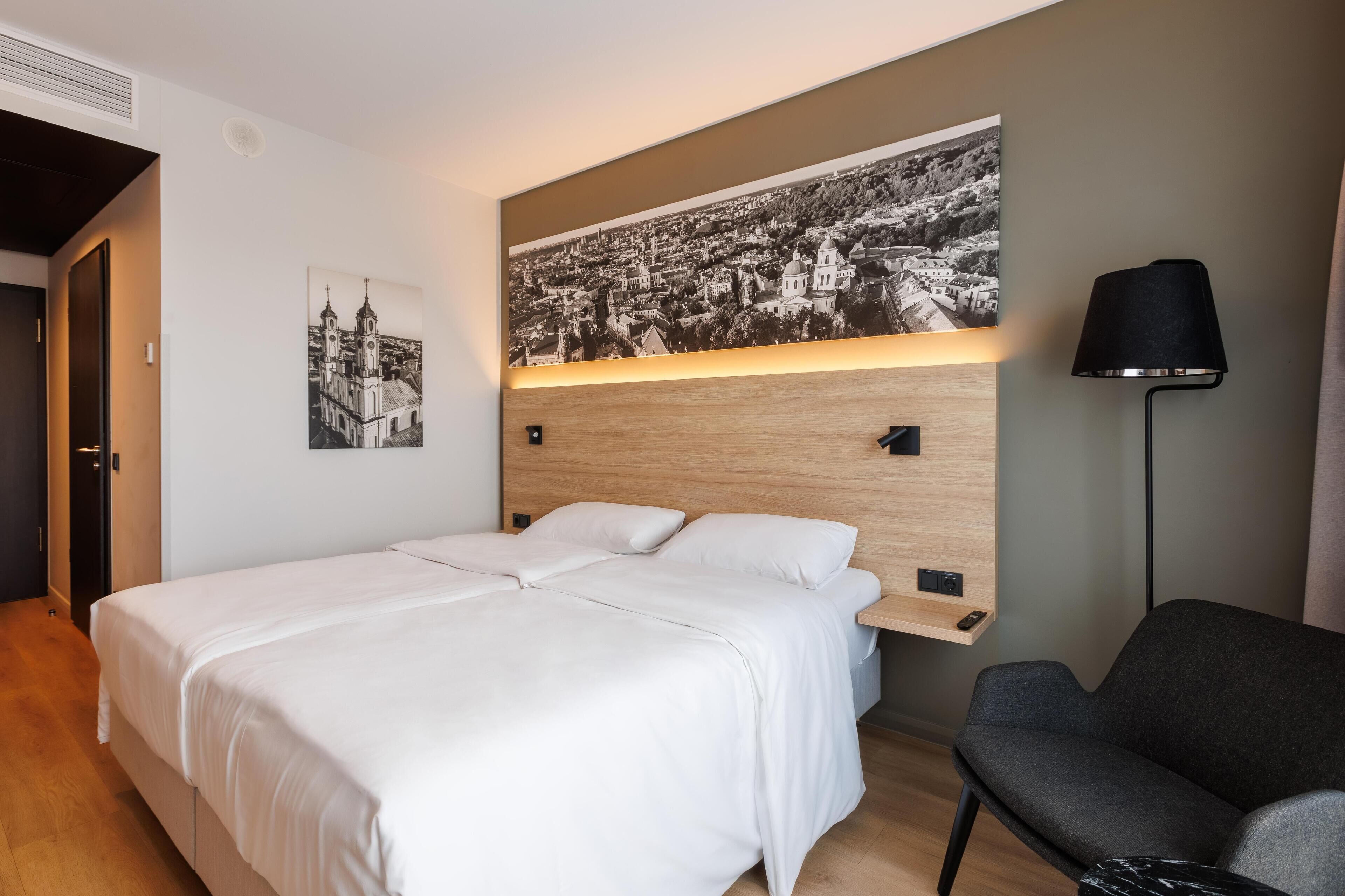 A hotel room with a bed, a desk, a chair, a lamp, and a wall-mounted shelf.