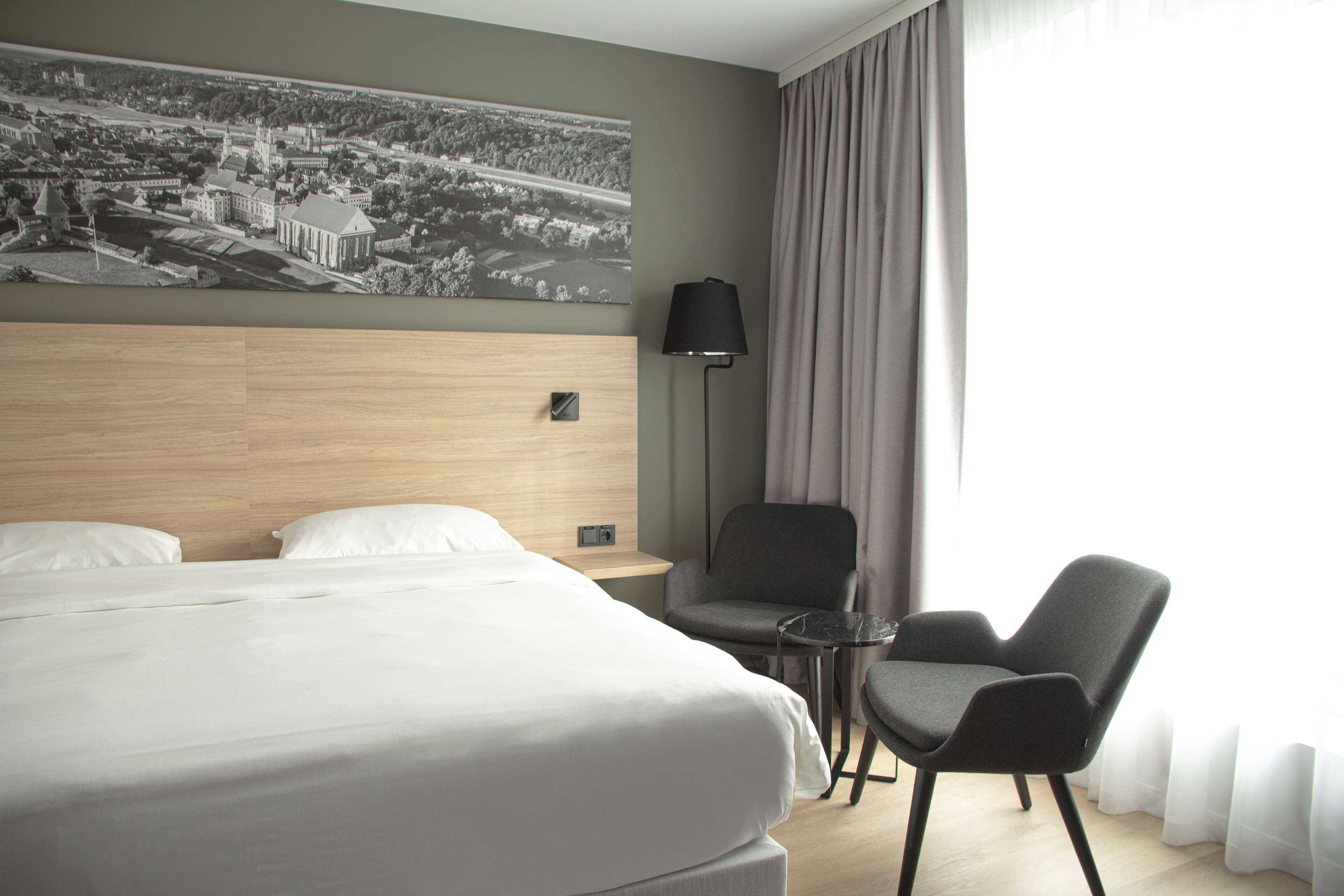 Habitación de hotel con una cama grande, dos sillones, una mesita, una lámpara y un cuadro de un paisaje urbano en la pared.