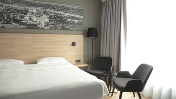 Habitación de hotel con una cama grande, dos sillones, una mesita, una lámpara y un cuadro de un paisaje urbano en la pared.