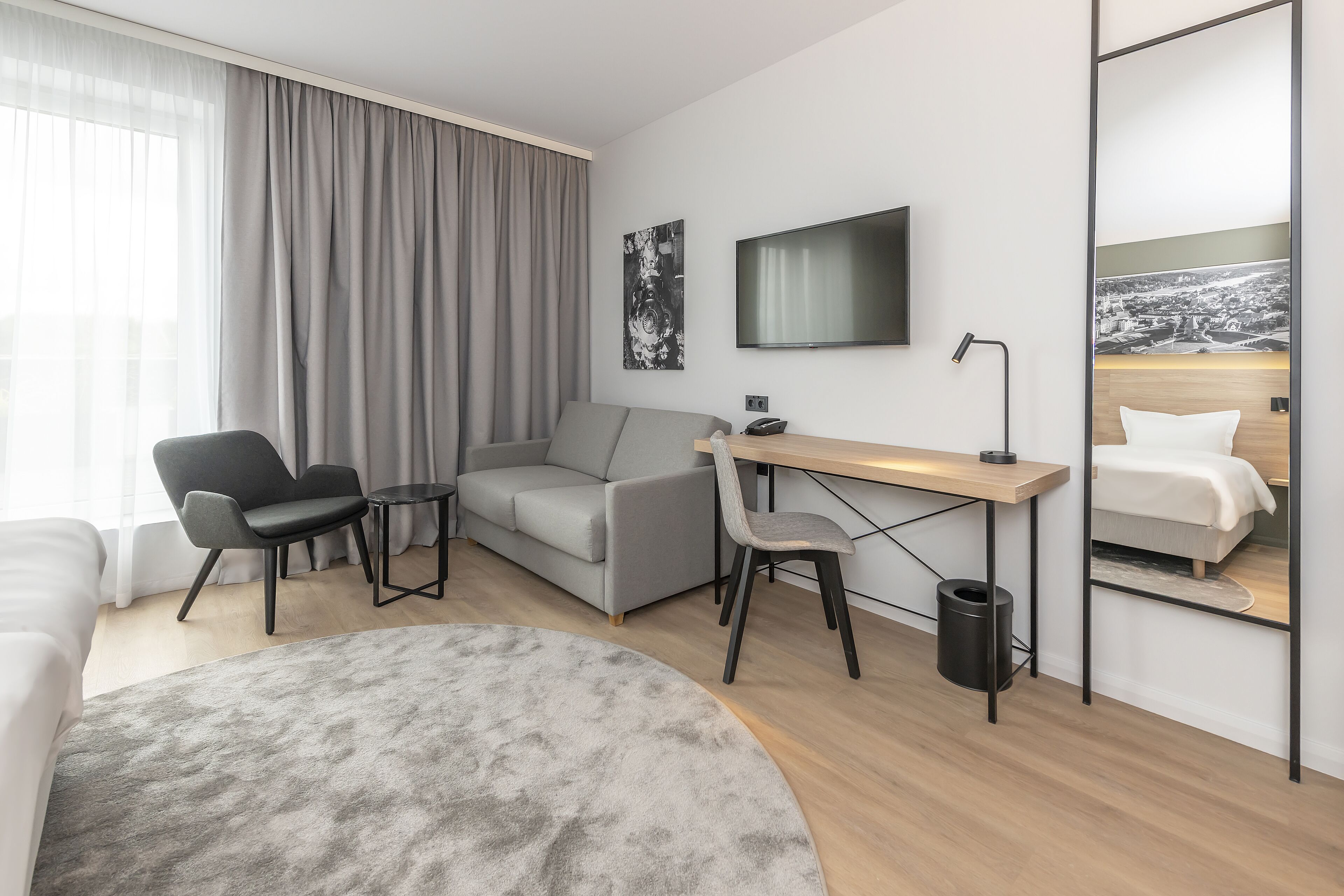 A modern hotel room with a grey sofa, a desk with a chair, a large mirror, and a flat-screen TV.