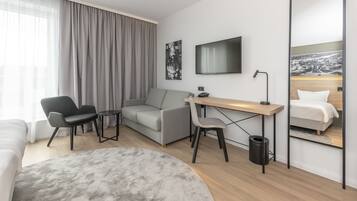 A modern hotel room with a grey sofa, a desk with a chair, a large mirror, and a flat-screen TV.
