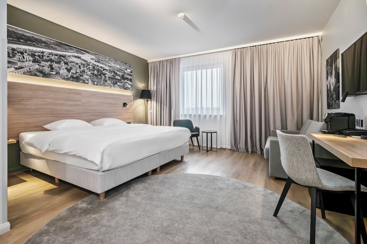 A modern hotel room with a large bed, a desk, a chair, a small table, and a large window with curtains.