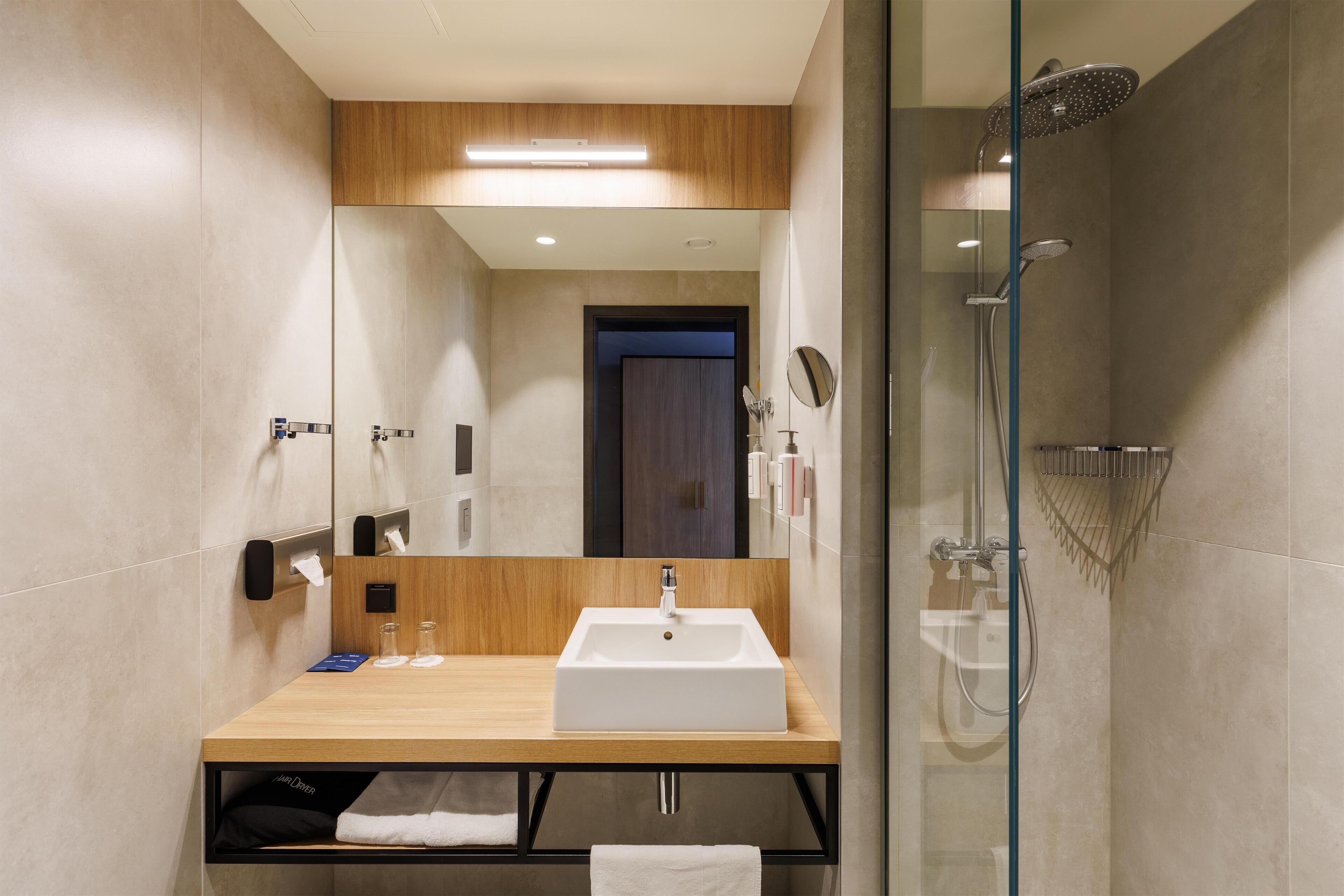 Un baño moderno con tocador de madera, un espejo grande, un lavamanos blanco y una zona de ducha con mampara de cristal.
