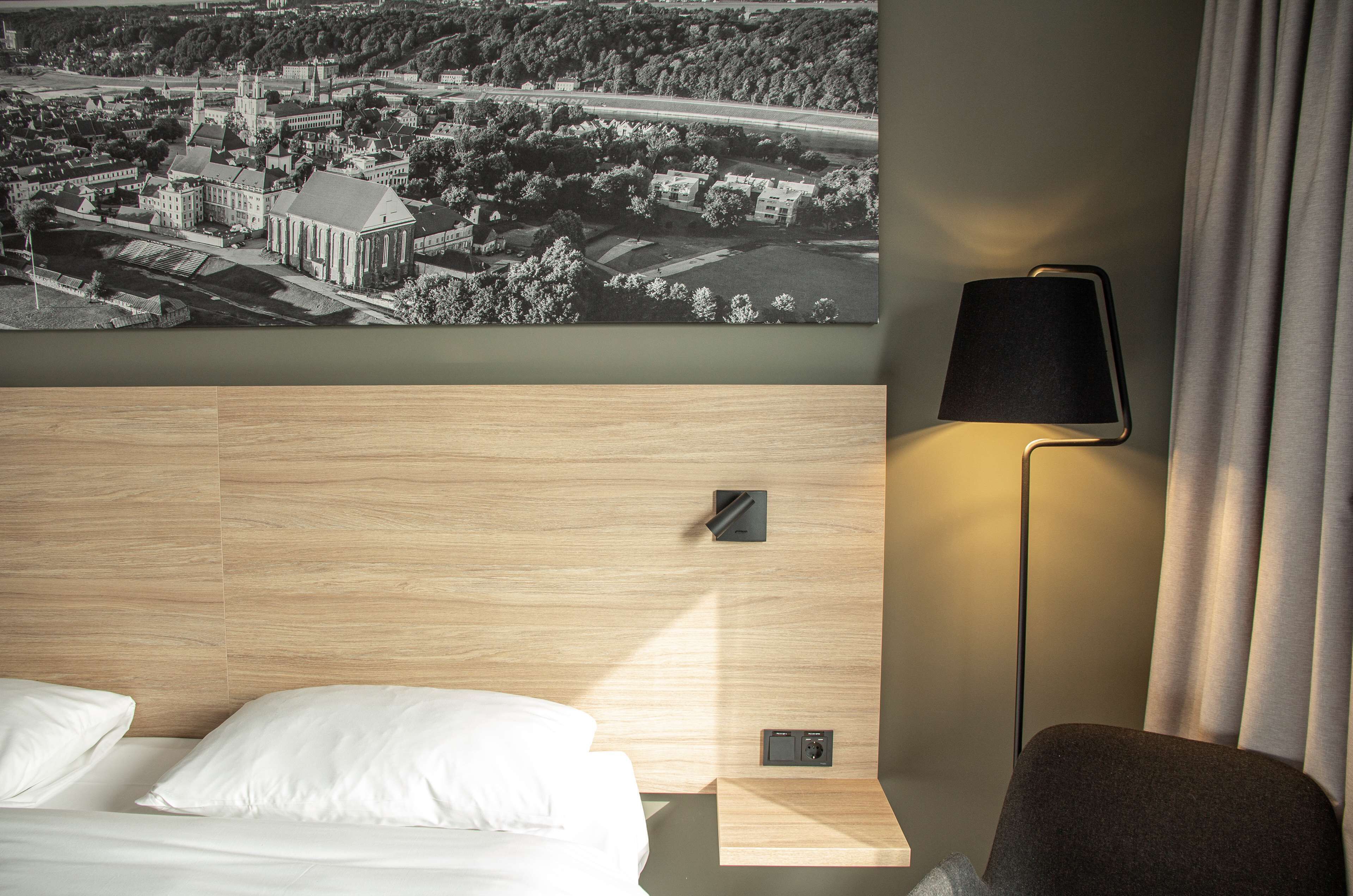 A hotel room with a bed, a nightstand, a lamp, and a large cityscape painting on the wall.