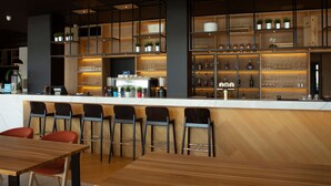 A modern bar area with a wooden counter, multiple seating options including bar stools and a table, and shelves stocked with bottles and glasses. - Park Inn by Radisson Vilnius Airport Hotel & Conference Centre (Vilnius)