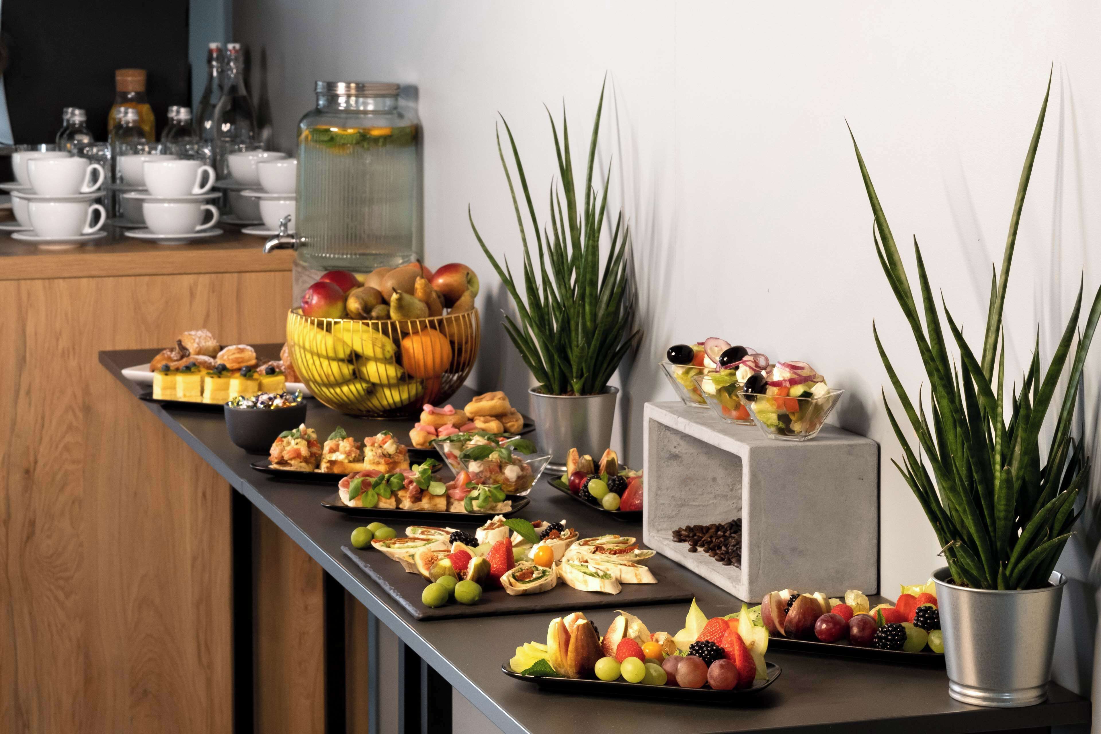 A buffet table with various food items and drinks, including fruits, pastries, and a large jar of pickles.