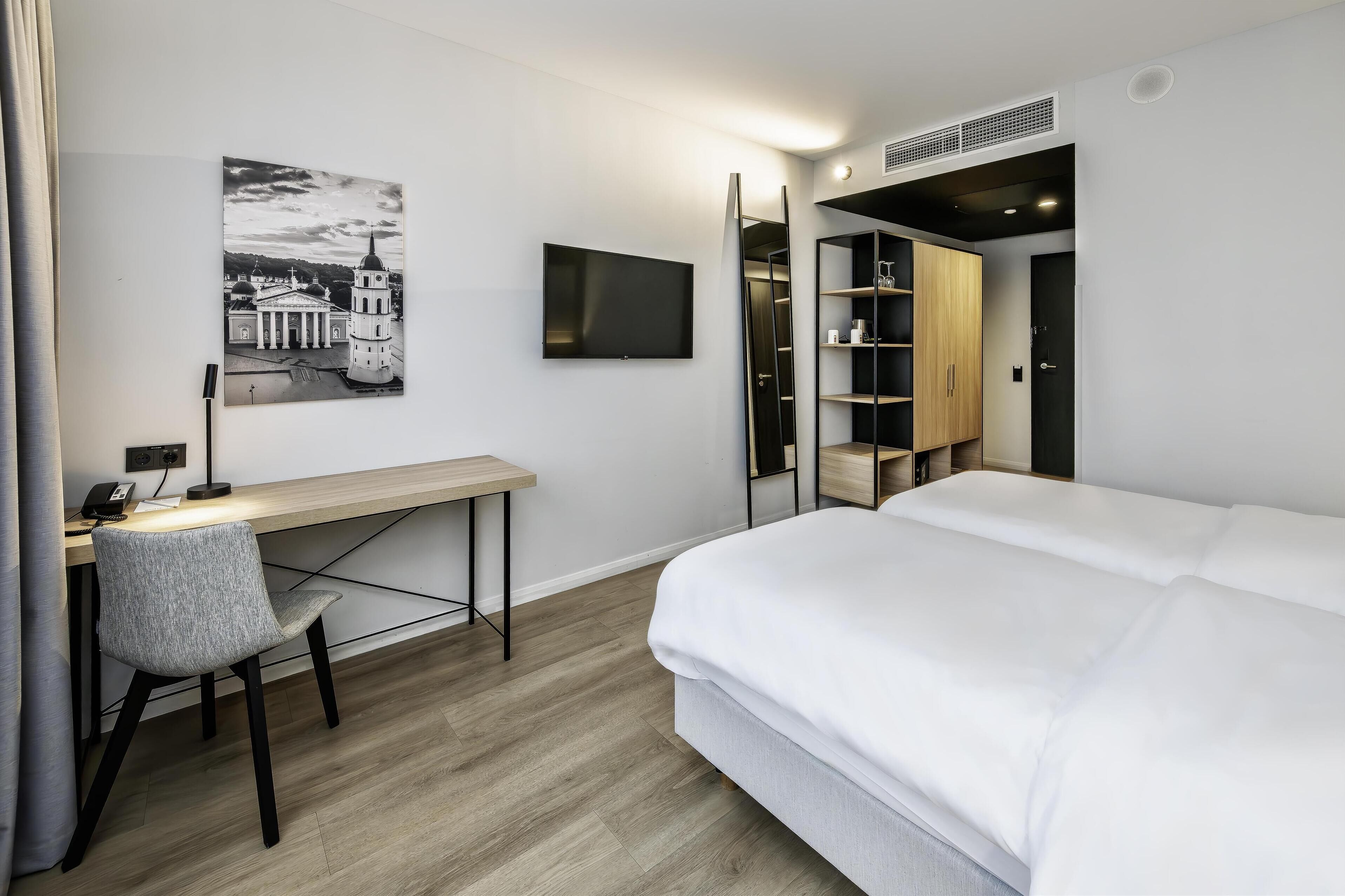A modern hotel room with a bed, a desk, a chair, a television, and a wardrobe.
