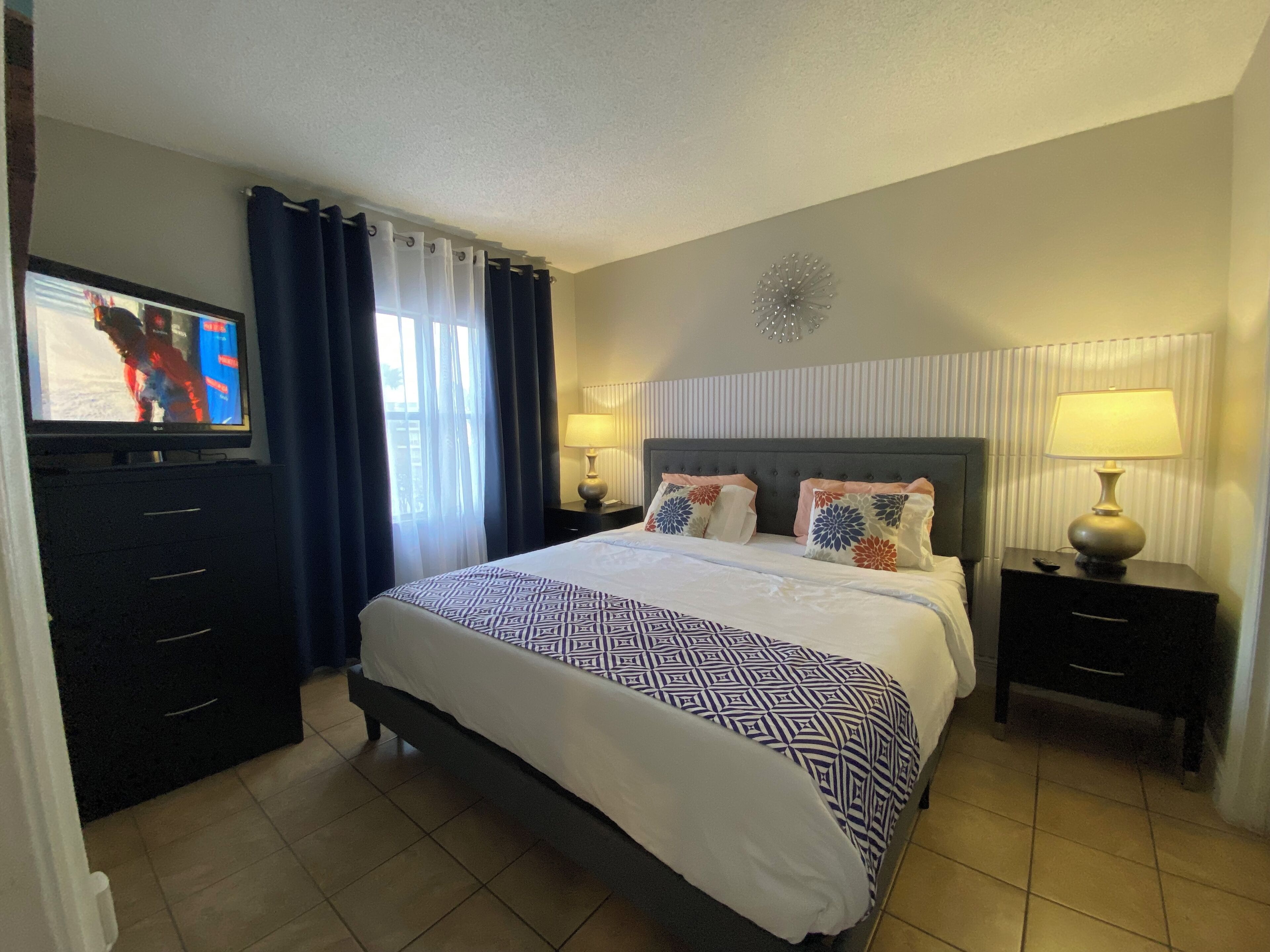 Een hotelkamer met een groot bed, twee nachtkastjes met lampen, een televisie op een dressoir en een raam met gordijnen.