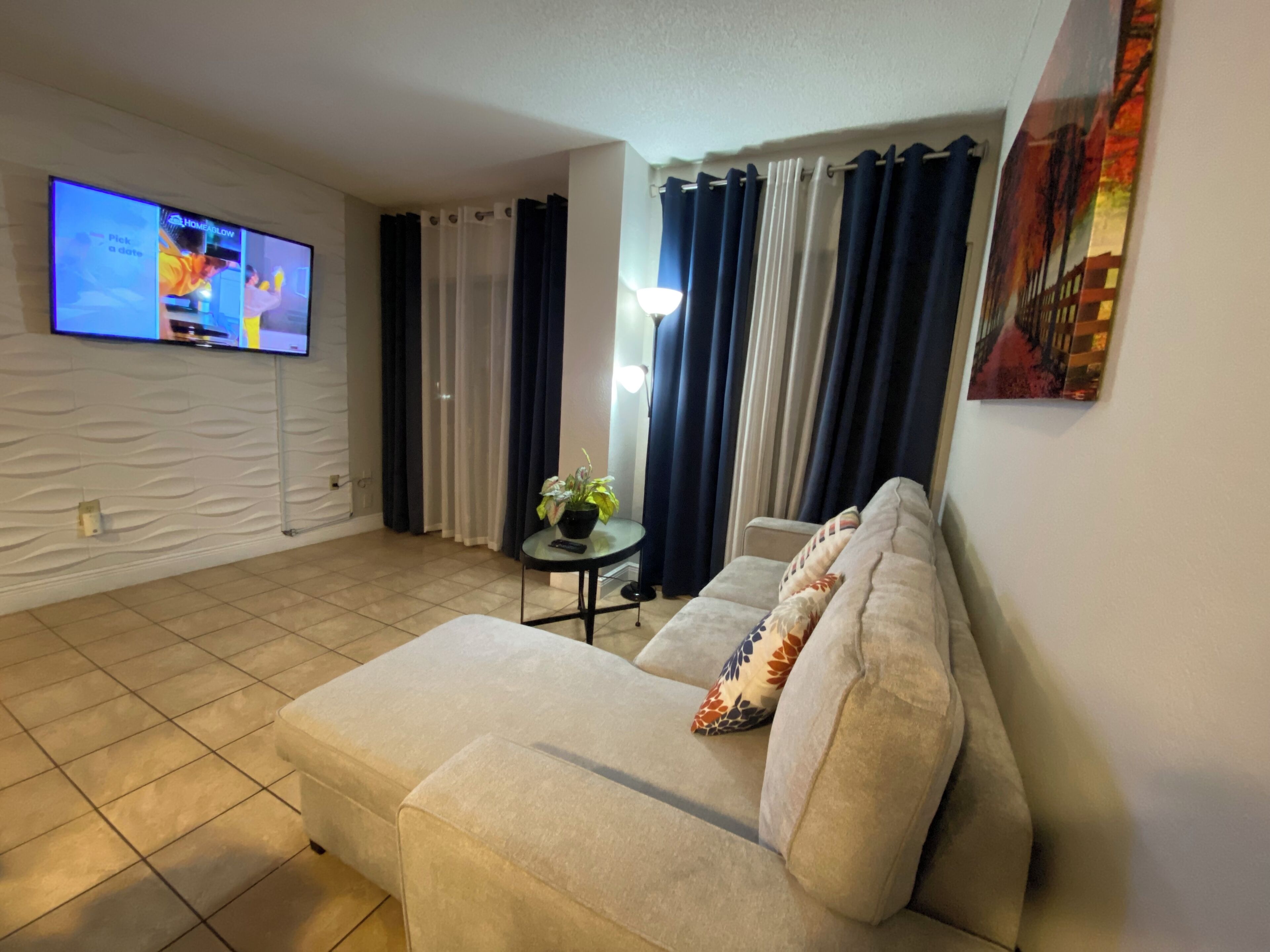 A living room with a beige sofa, a small table with a plant, a TV mounted on the wall, and a painting on the wall.