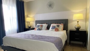 A neatly made bed with a patterned bedspread, two bedside tables with lamps, a decorative wall piece, and a window with curtains. - 2 BR RESORT IN PERFECT LOCATION NEAR UNIVERSAL (Orlando)