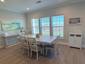 A dining area with a wooden table and chairs, large windows with blinds, a painting on the wall, and a cabinet with glass doors.