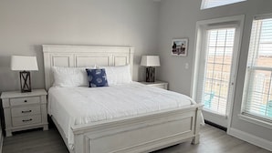 Un dormitorio con una cama blanca, dos mesitas de noche con lámparas, un ventilador de techo y un ventanal con persianas.