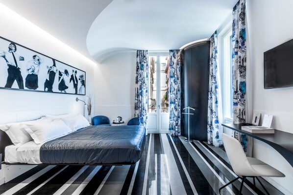 A modern hotel room with a bed, a desk, and a chair. There is a painting on the wall and a window with curtains.