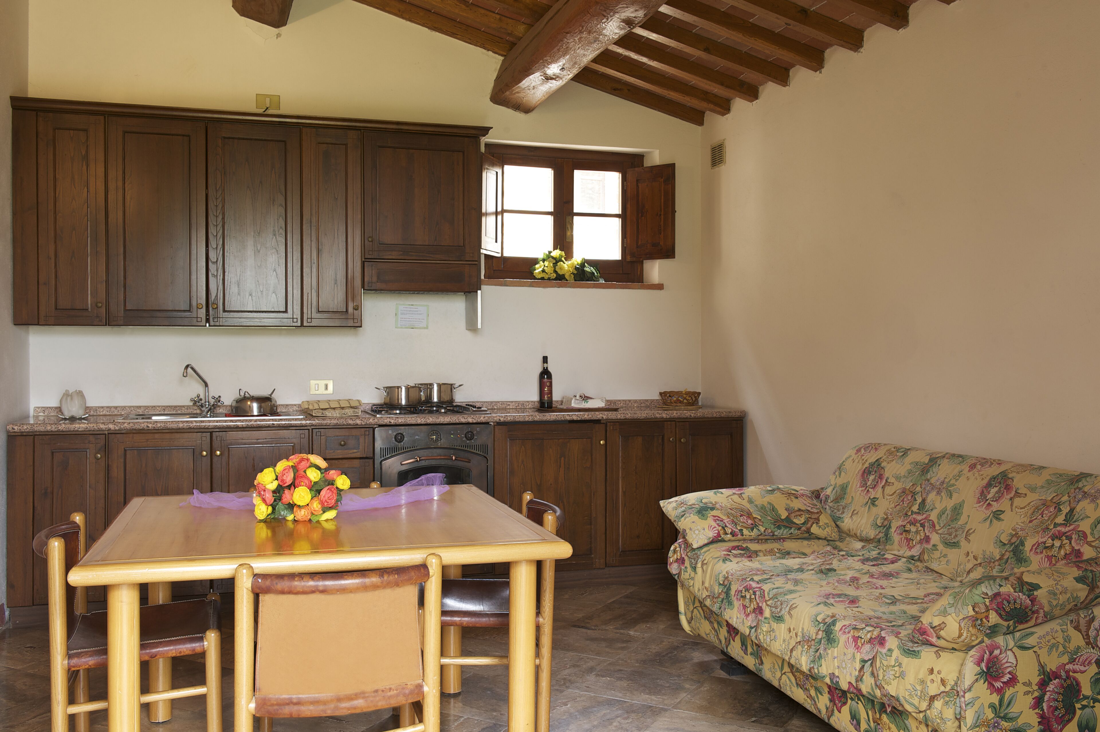 Una cocina con armarios de madera, una mesa de comedor con sillas, un sofá floral y una ventana con flores.