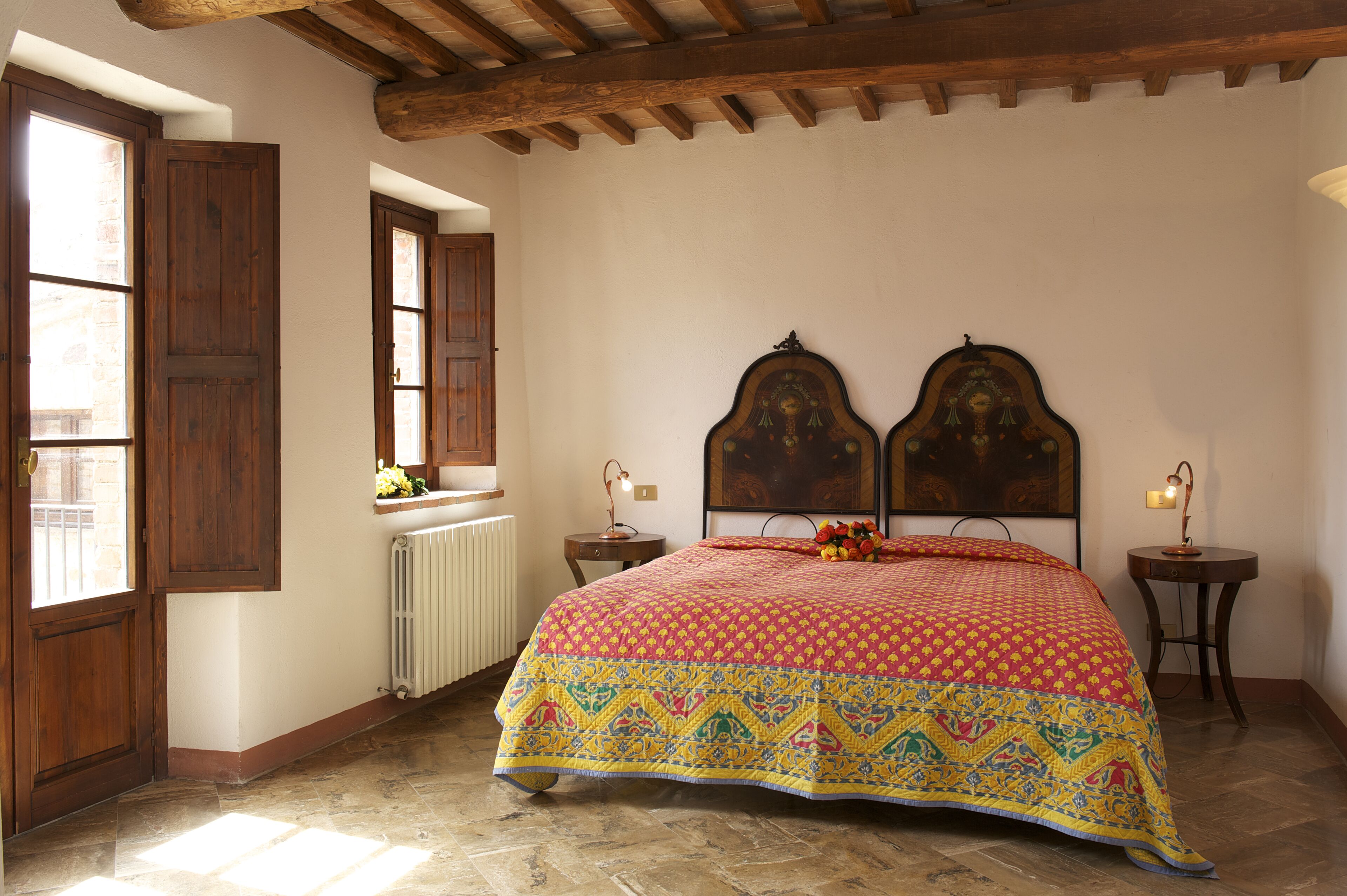Een slaapkamer met een groot bed, houten hoofdbord, een raam met uitzicht, een radiator en een klein tafeltje met een lamp.